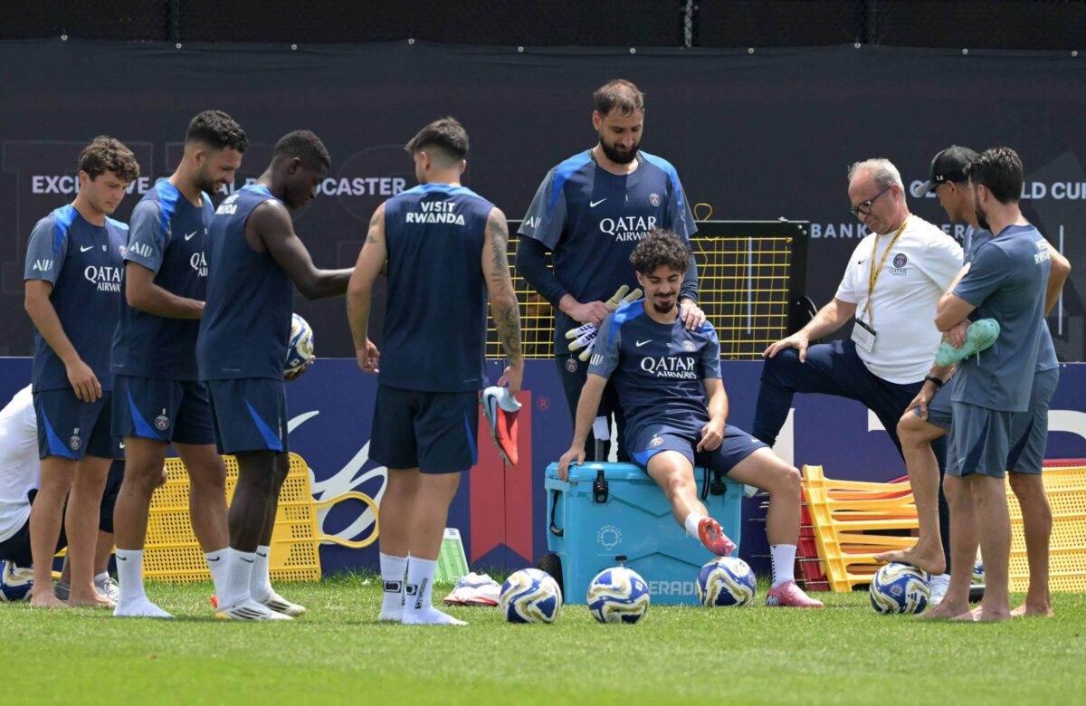 Um grupo de jogadores de futebol está reunido em um campo durante um treino. Eles vestem uniformes azuis e brancos, com alguns jogadores em pé e outros sentados. Um dos jogadores está sentado em um banco, enquanto um treinador ou membro da comissão técnica está ao seu lado, conversando. Há bolas de futebol no chão e um fundo com banners e uma rede de proteção.