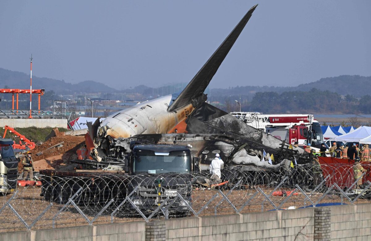 A imagem mostra os destroços de um avião acidentado em um local de pouso. A fuselagem do avião está danificada e parcialmente queimada, com a asa visivelmente quebrada. Há veículos de emergência e equipes de resgate ao redor, além de uma cerca de arame farpado ao fundo. O céu está claro e há montanhas ao longe.