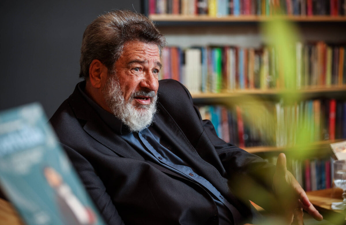Um homem com cabelo grisalho e barba espessa, vestido com um paletó escuro e uma camisa azul, está sentado em um ambiente interno. Ao fundo, há uma estante repleta de livros. O homem parece estar em uma conversa, gesticulando com as mãos. À frente dele, há uma planta e um copo de água sobre uma mesa.