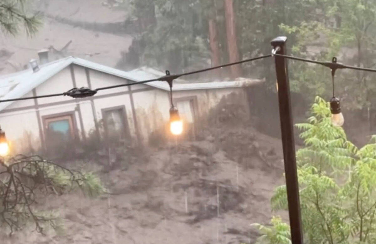 A imagem mostra uma cena de um deslizamento de terra em meio a uma forte chuva. É possível ver uma área de terra e vegetação sendo arrastada, com uma casa visível ao fundo. A visibilidade é baixa devido à chuva intensa, e há uma estrutura de madeira em primeiro plano, com luzes penduradas. A vegetação ao redor parece densa e verde.