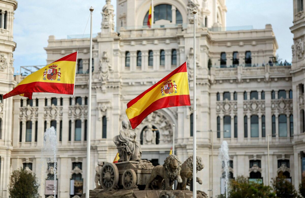 A imagem mostra uma praça com duas bandeiras da Espanha hasteadas em postes. Ao fundo, há um edifício imponente, que parece ser um palácio, com detalhes arquitetônicos elaborados. Na frente, há uma fonte e uma escultura de uma figura mitológica em uma carruagem, cercada por leões. O céu está claro e a atmosfera é de um dia ensolarado.