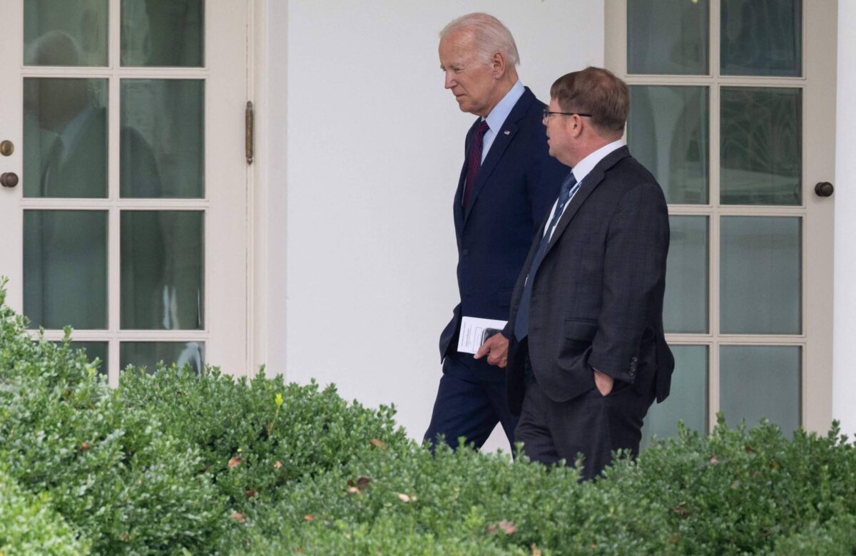 Na imagem, duas pessoas estão caminhando lado a lado em um ambiente externo, possivelmente no jardim da Casa Branca. A pessoa à esquerda está vestida com um terno escuro e gravata, enquanto a pessoa à direita usa um terno cinza e óculos. Ao fundo, há portas de vidro e uma vegetação baixa na frente.
