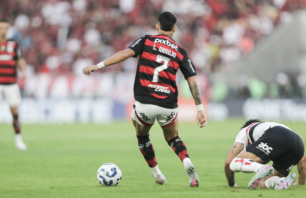 A imagem mostra um jogador de futebol com a camisa do Flamengo, número 7, correndo em direção à bola. Ele está de costas e se afasta de um adversário que está caindo no chão. O fundo é um estádio com torcedores visíveis, criando um ambiente de jogo intenso.