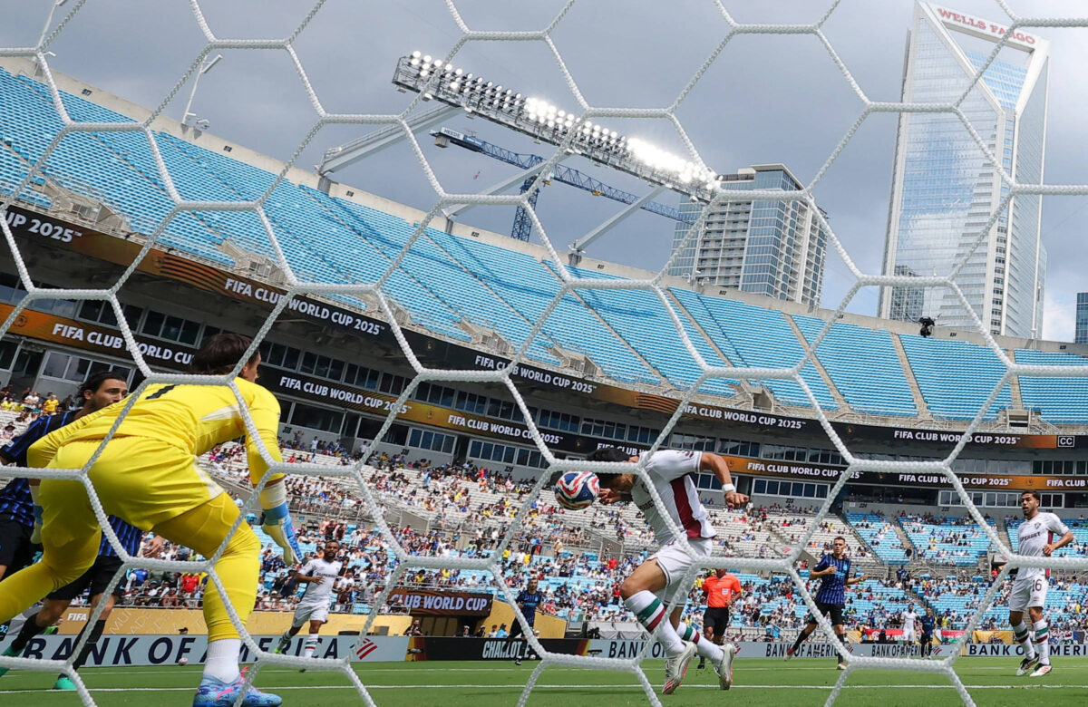 A imagem mostra um jogo de futebol em um estádio. Um jogador está prestes a chutar a bola em direção ao gol, enquanto o goleiro, vestido de amarelo, se prepara para defender. A rede do gol está em primeiro plano, e ao fundo, é possível ver as arquibancadas com torcedores e a estrutura do estádio. O céu está parcialmente nublado e há prédios altos ao fundo.