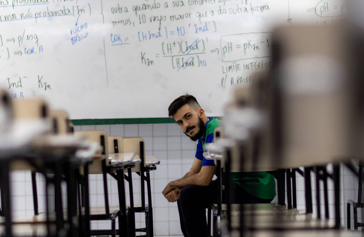 Um jovem sentado no chão de uma sala de aula, entre as mesas e cadeiras. Ele tem cabelo curto e barba, vestindo uma camiseta azul e um colete verde. Ao fundo, uma lousa branca com anotações e fórmulas escritas em caneta preta e azul. A sala parece vazia, com as mesas organizadas em fileiras.