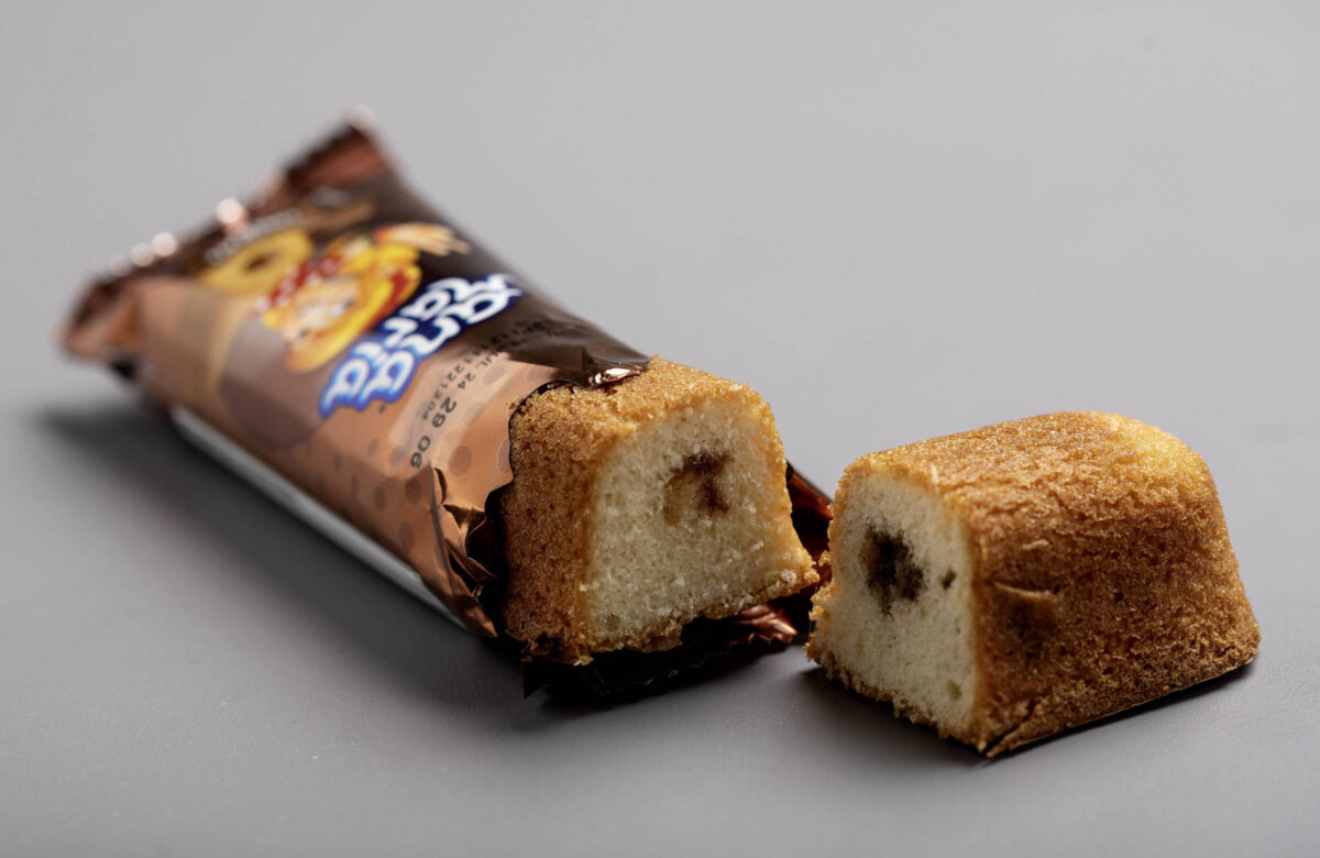 A imagem mostra um bolo de chocolate cortado ao meio, revelando um recheio de chocolate. O bolo é de cor clara com uma cobertura marrom. Ao fundo, há uma embalagem de papel que contém o bolo, com a parte superior visível, onde se pode ler o nome do produto. O fundo da imagem é cinza.