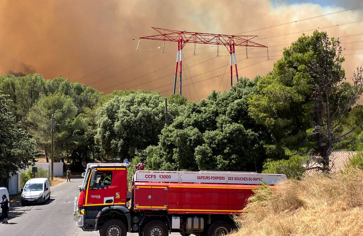 A imagem mostra um caminhão de bombeiros estacionado em uma rua, com vegetação densa ao redor. Ao fundo, há uma grande nuvem de fumaça laranja, indicando um incêndio florestal. Estruturas de energia elétrica são visíveis, com torres de transmissão se destacando contra o céu. O cenário é de emergência, com a presença de árvores e um terreno seco.