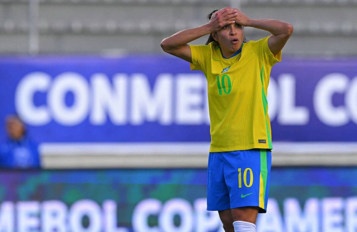 A imagem mostra uma jogadora de futebol em um campo, vestindo uma camisa amarela com detalhes em verde, número 10, e calças brancas, com as mãos na cabeça, aparentando frustração. Ao fundo, há uma faixa com o texto