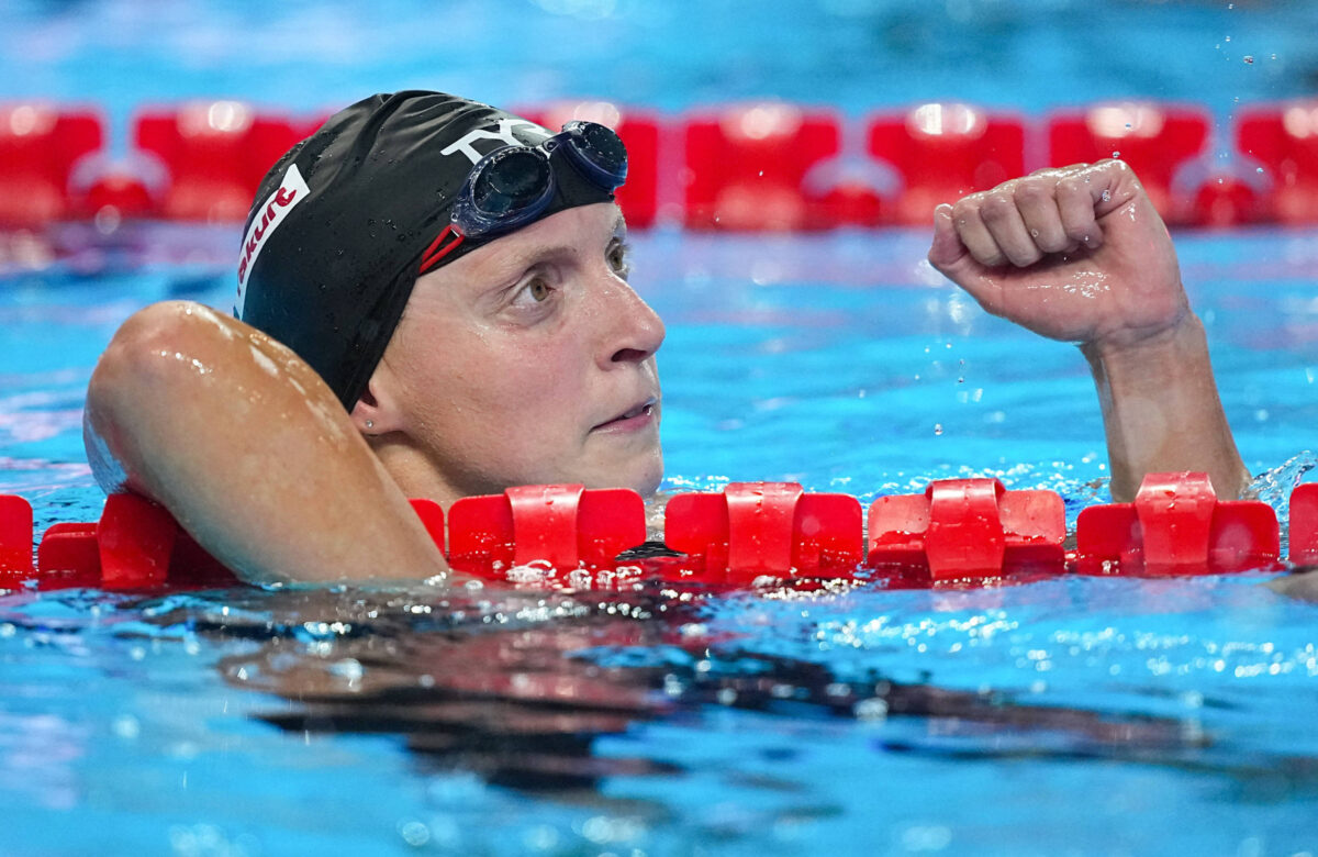 A imagem mostra uma nadadora em uma piscina, com a cabeça acima da água e apoiada na borda da piscina. Ela está usando um boné de natação preto e óculos de natação. A nadadora parece estar em um momento de reflexão ou celebração, levantando o braço direito com o punho cerrado. O fundo da imagem é composto por água azul e as bordas da piscina são marcadas por flutuadores vermelhos.