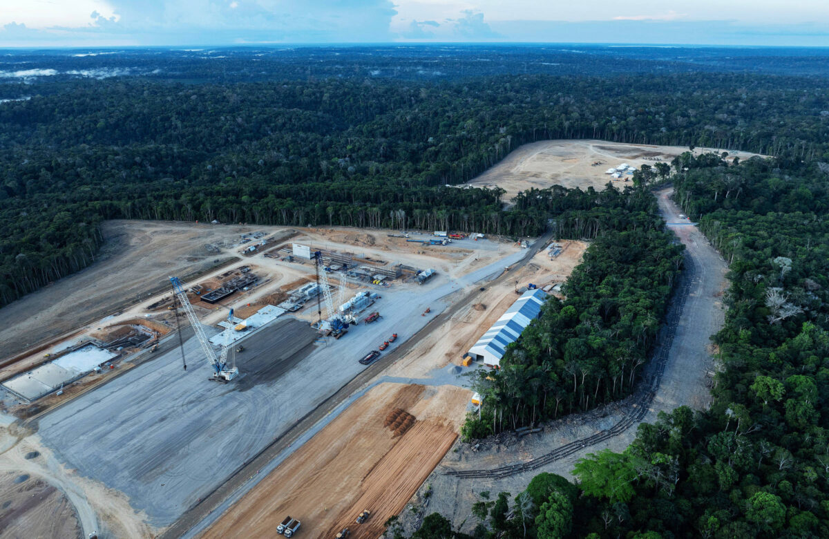 A imagem mostra uma área de construção em uma região florestal. No primeiro plano, há uma grande área desmatada com maquinário e estruturas de construção visíveis. Ao fundo, a floresta densa se estende até o horizonte, com árvores altas e uma paisagem verde. O céu está parcialmente nublado, e há uma estrada que se afasta da área de construção.