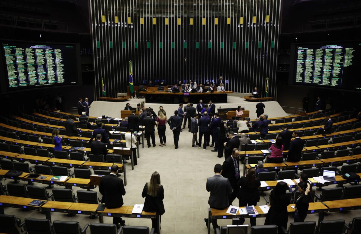 A imagem mostra o plenário da Câmara dos Deputados do Brasil, com várias pessoas em pé e sentadas. Ao fundo, há uma mesa com representantes e bandeiras do Brasil. As paredes laterais estão equipadas com painéis eletrônicos que exibem informações. O ambiente é amplo, com cadeiras dispostas em fileiras e um piso claro.