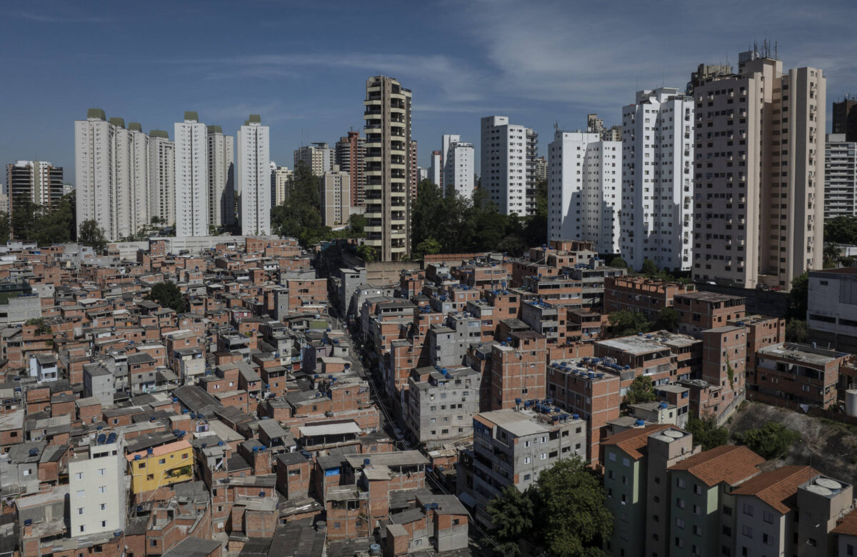 A imagem mostra uma vista panorâmica de uma área urbana, onde à esquerda há uma densa aglomeração de casas simples e coloridas, típicas de uma comunidade de baixa renda. À direita, destacam-se altos edifícios modernos e brancos, representando uma parte mais desenvolvida da cidade. O céu está limpo e azul, e a vegetação é visível entre as construções.