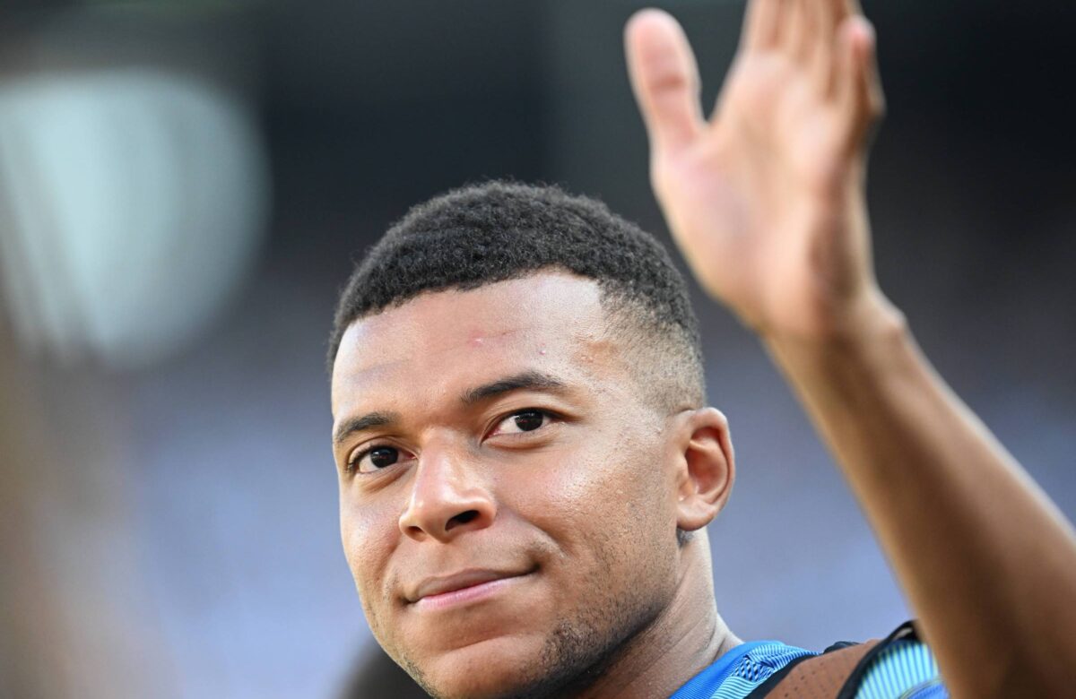 Um jogador de futebol está acenando com a mão. Ele tem cabelo curto e usa uma camisa esportiva azul. O fundo é desfocado, sugerindo que ele está em um estádio ou campo de futebol.