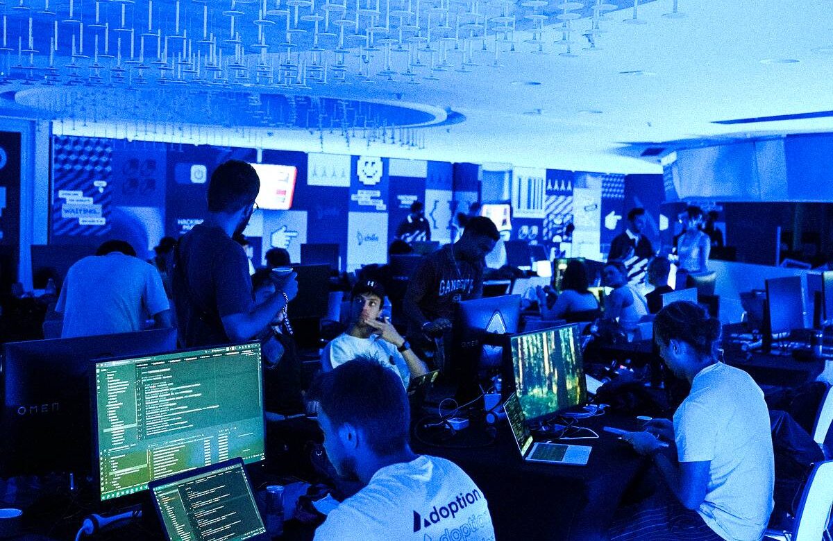 A imagem mostra um ambiente de trabalho com várias pessoas em um espaço iluminado em tom azul. Há várias mesas com computadores, e algumas pessoas estão sentadas, enquanto outras estão em pé. As telas dos computadores exibem códigos e gráficos. O ambiente parece ser um evento ou uma competição, com banners e logotipos visíveis nas paredes ao fundo.