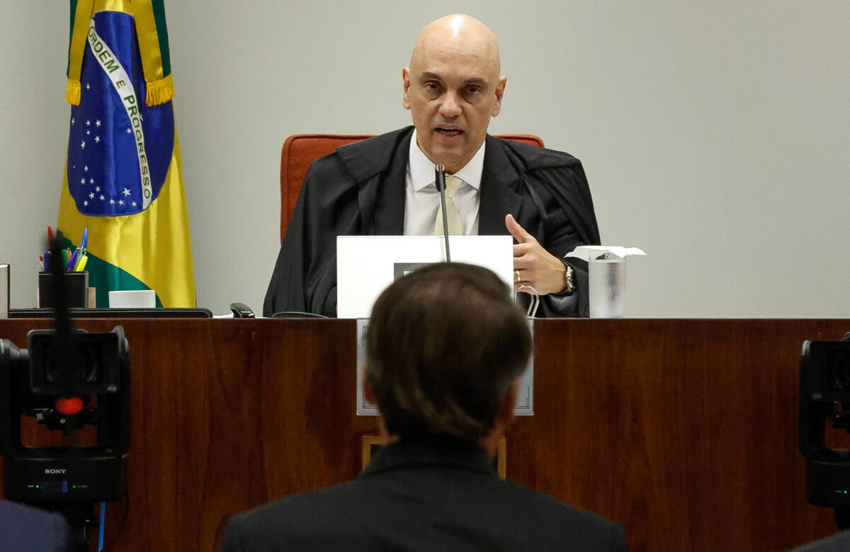 A imagem mostra um juiz em uma audiência, vestido com uma toga preta e uma camisa branca, sentado em uma mesa de tribunal. Ele está gesticulando enquanto fala, com um copo de água e canetas ao seu lado. Ao fundo, há uma bandeira do Brasil. Na frente do juiz, há uma pessoa de costas, aparentemente ouvindo atentamente.