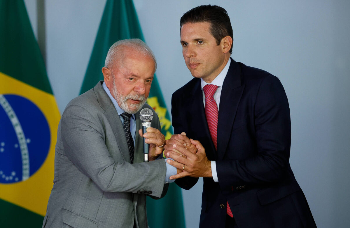 Na imagem, dois homens estão em um evento oficial. O homem à esquerda, com cabelo grisalho e vestindo um terno cinza, segura um microfone e parece estar falando. O homem à direita, com cabelo escuro e vestindo um terno escuro, está segurando a mão do homem à esquerda, olhando para ele. Ao fundo, há bandeiras do Brasil.
