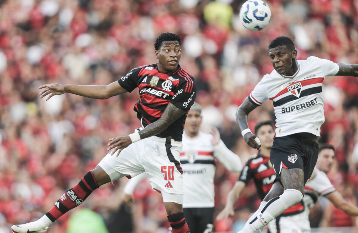 A imagem mostra um momento de um jogo de futebol entre as equipes do Flamengo e do São Paulo. Um jogador do Flamengo, vestindo uma camisa listrada em vermelho e preto, está saltando para interceptar a bola, enquanto um jogador do São Paulo, vestido com uma camisa branca e detalhes em vermelho e preto, também está em ação. Ao fundo, é possível ver outros jogadores e a torcida nas arquibancadas, que estão predominantemente vestidas com as cores do Flamengo.