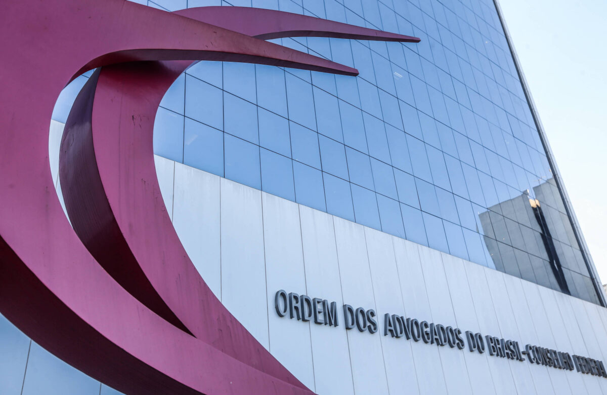 A imagem mostra a fachada de um edifício moderno com uma grande escultura em forma de ondas vermelhas na parte superior esquerda. Na parte inferior direita, está escrito em letras grandes e maiúsculas: