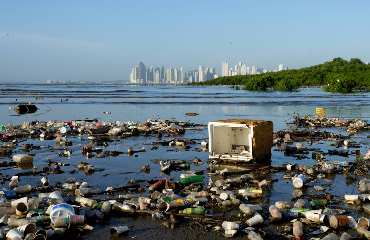 A imagem mostra uma área costeira poluída, com uma grande quantidade de lixo, incluindo copos plásticos, em meio à água. Ao fundo, é possível ver uma cidade com arranha-céus e vegetação ao longo da costa.