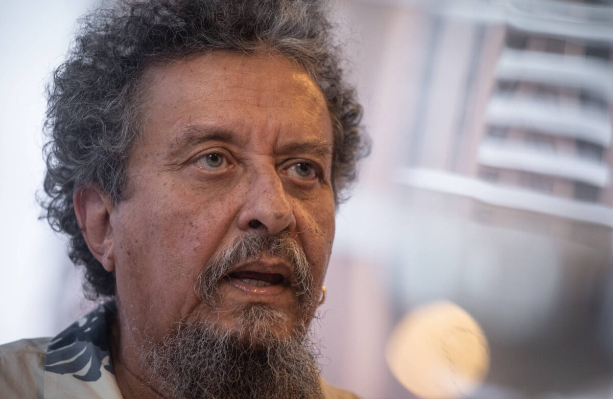 Um homem com cabelo grisalho e barba, usando uma camisa de estampas claras. Ele está em um ambiente interno, com um fundo desfocado que sugere um espaço urbano. O homem parece estar em um momento de reflexão ou fala, com uma expressão séria.