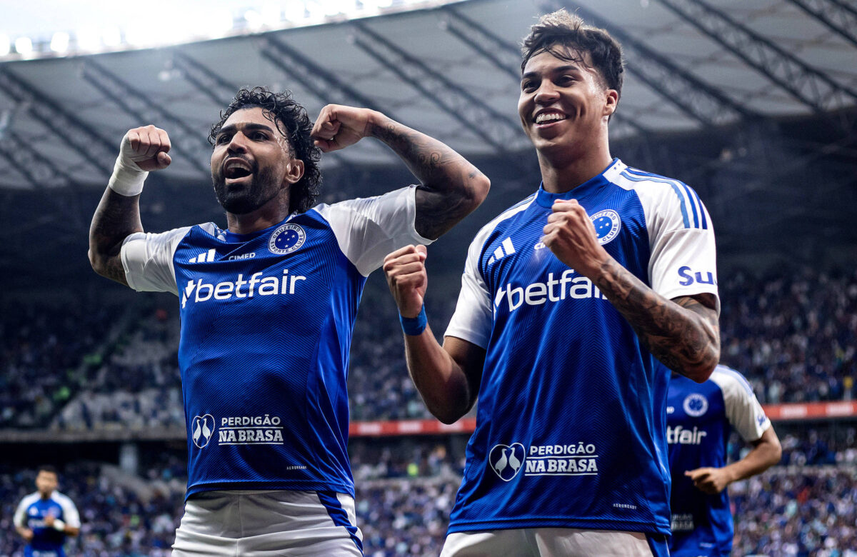 Dois jogadores de futebol estão comemorando em campo. Ambos vestem uniformes azuis com detalhes brancos e logotipos. O jogador à esquerda levanta os braços em celebração, enquanto o jogador à direita sorri e também levanta o braço. Ao fundo, há uma multidão de torcedores em um estádio.