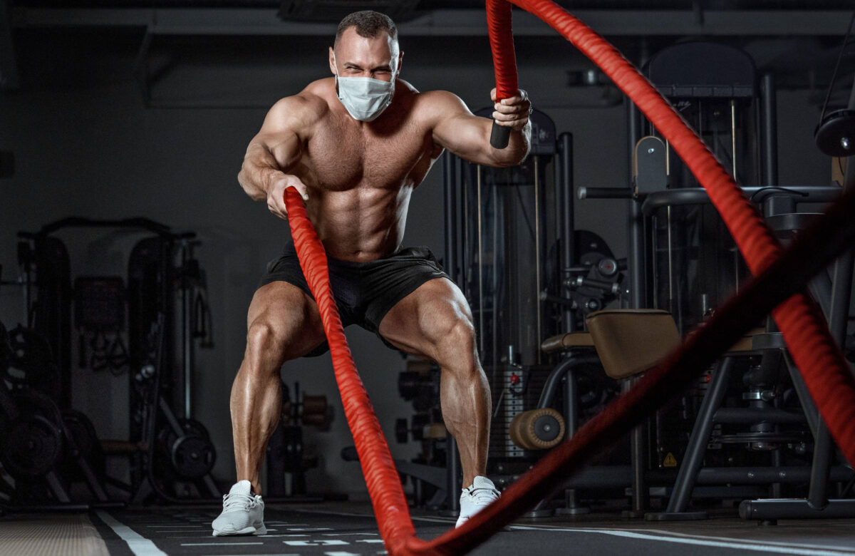 Um homem musculoso está em uma academia, usando uma máscara facial. Ele está em posição de agachamento, segurando cordas de batalha vermelhas, puxando-as para cima e para baixo. O ambiente é bem iluminado, com equipamentos de musculação ao fundo.