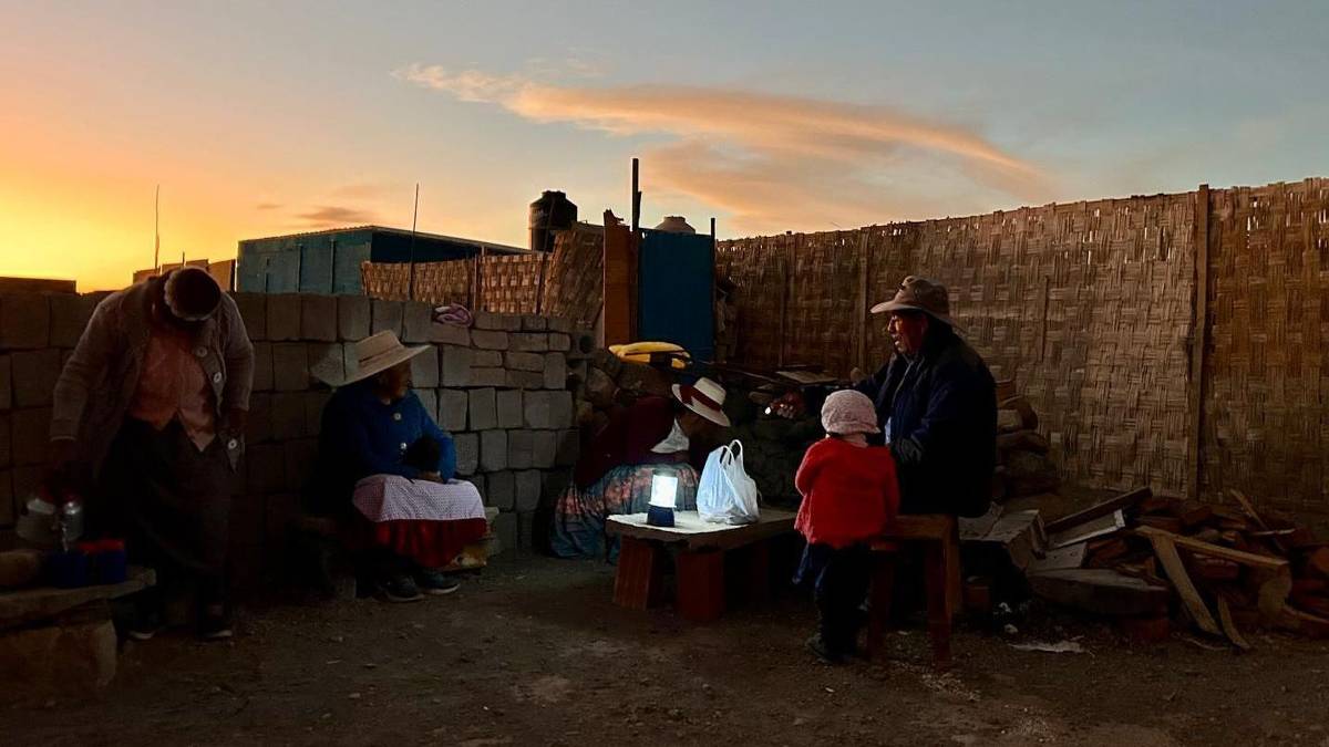 A imagem mostra um grupo de pessoas sentadas em um ambiente rústico ao entardecer. Há uma mesa baixa com uma luz em cima, iluminando o espaço. As pessoas estão vestidas com roupas tradicionais, algumas usando chapéus. O fundo é composto por paredes de tijolos e uma cerca de bambu, com um céu colorido ao fundo.