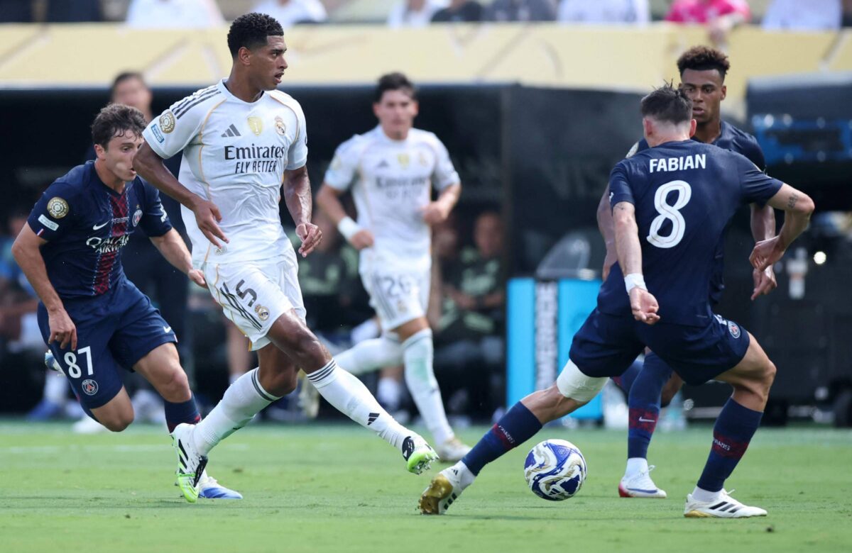 A imagem mostra um momento de um jogo de futebol entre duas equipes. Um jogador do time da esquerda, vestindo uma camisa azul com detalhes vermelhos, está se aproximando de um jogador do time da direita, que está usando uma camisa branca com detalhes dourados. O jogador da camisa branca parece estar driblando a bola, enquanto outro jogador do time da esquerda, também em azul, observa a jogada. Ao fundo, há outros jogadores e uma arquibancada com torcedores.