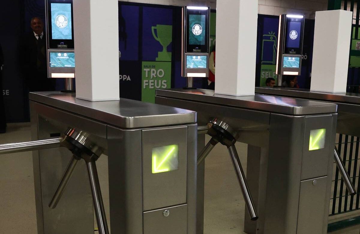 A imagem mostra várias catracas de acesso em um ambiente interno. As catracas são de metal e possuem um design moderno, com um painel digital em cada uma, exibindo informações. Ao fundo, há uma parede com gráficos e textos, incluindo a palavra