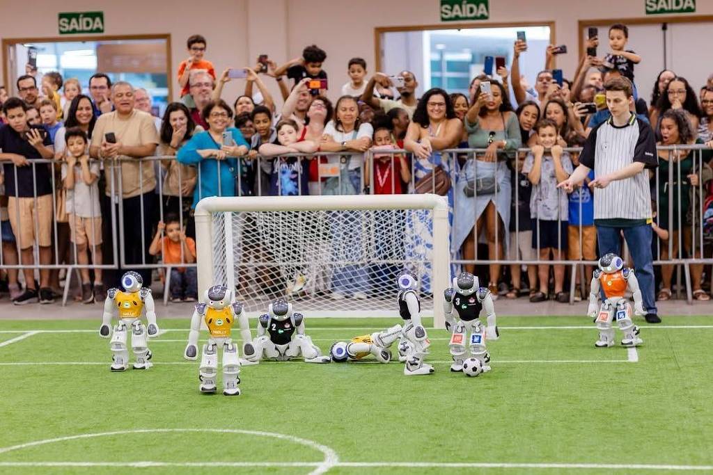 A imagem mostra um campo de futebol com robôs jogando. Há seis robôs em campo, alguns com uniformes coloridos, e um deles está caído no chão. Ao fundo, uma multidão de pessoas assiste ao jogo, algumas filmando com celulares. O gol é visível, e há uma rede atrás dele. O ambiente parece ser um evento ou competição de robôs.
