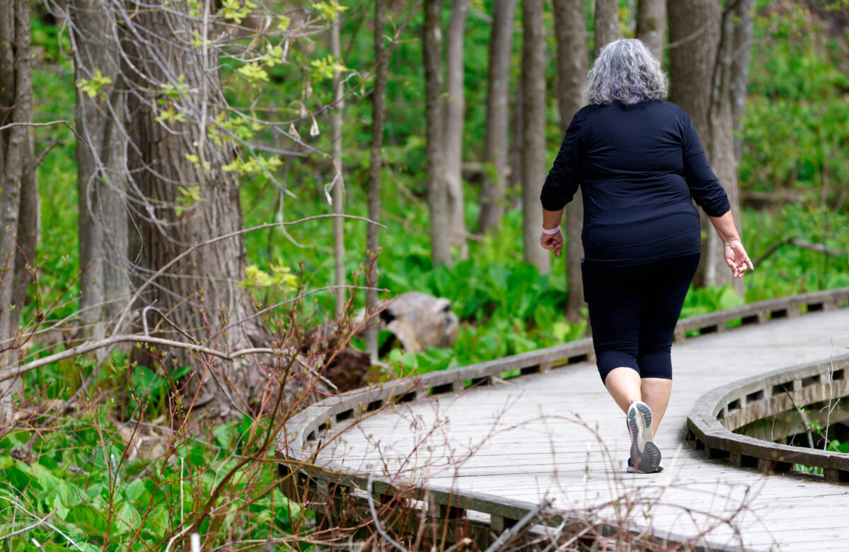 Uma mulher com cabelo grisalho está caminhando por uma trilha de madeira em uma área verde. Ela usa uma camiseta preta e calças legging pretas. Ao fundo, há árvores e vegetação densa, criando um ambiente natural e tranquilo.