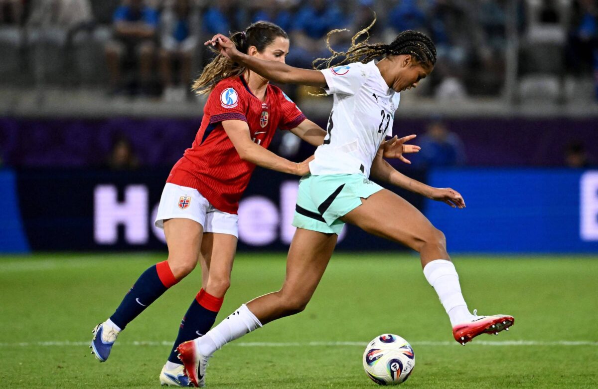 A imagem mostra duas jogadoras de futebol em um campo. A jogadora à esquerda, vestindo uma camisa vermelha e calças escuras, está tentando alcançar a jogadora à direita, que está vestindo uma camisa branca e calças claras. A jogadora da direita está em movimento, prestes a chutar a bola que está no chão. O fundo apresenta um estádio com espectadores.