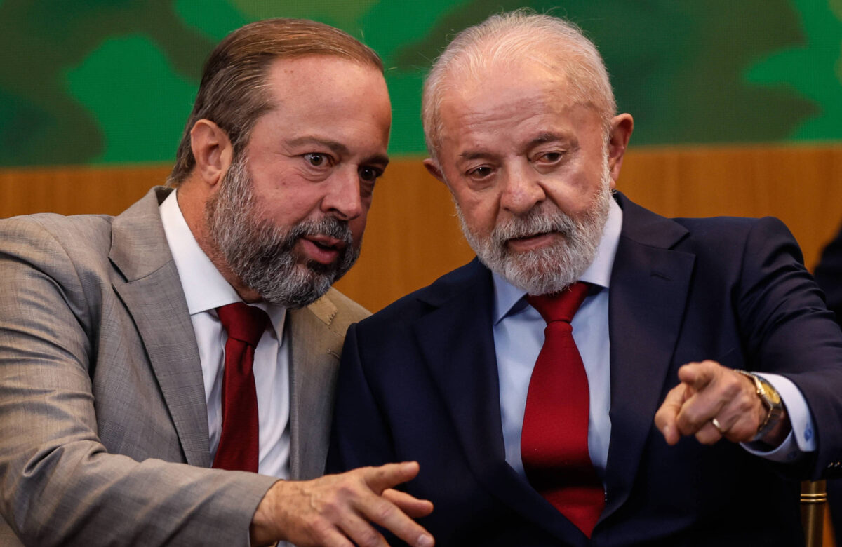 Na imagem, dois homens estão sentados próximos um do outro, em um ambiente formal. O homem à esquerda, com cabelo castanho e barba, veste um terno cinza claro e uma gravata vermelha. O homem à direita, com cabelo grisalho e barba, usa um terno escuro e uma gravata vermelha. Eles parecem estar em uma conversa íntima, com o homem à direita apontando para algo fora do quadro.