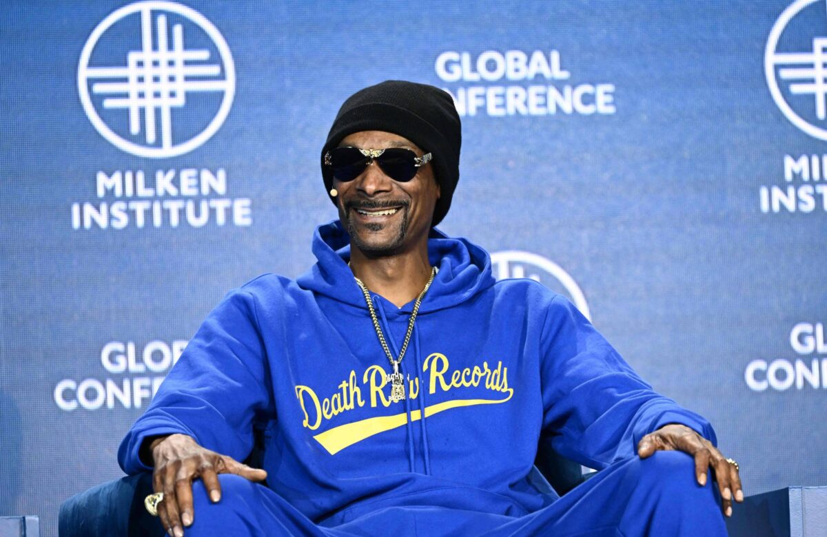 Um homem sorridente está sentado em uma cadeira em um evento. Ele usa um moletom azul com a inscrição