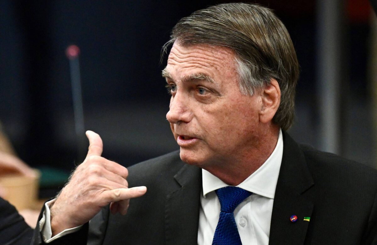 Um homem com cabelo castanho e liso, usando um terno preto e uma gravata azul, está em uma reunião. Ele está olhando para o lado e fazendo um gesto com a mão, como se estivesse fazendo um sinal de telefone. Ao fundo, há uma mesa e outros elementos de um ambiente de reunião.