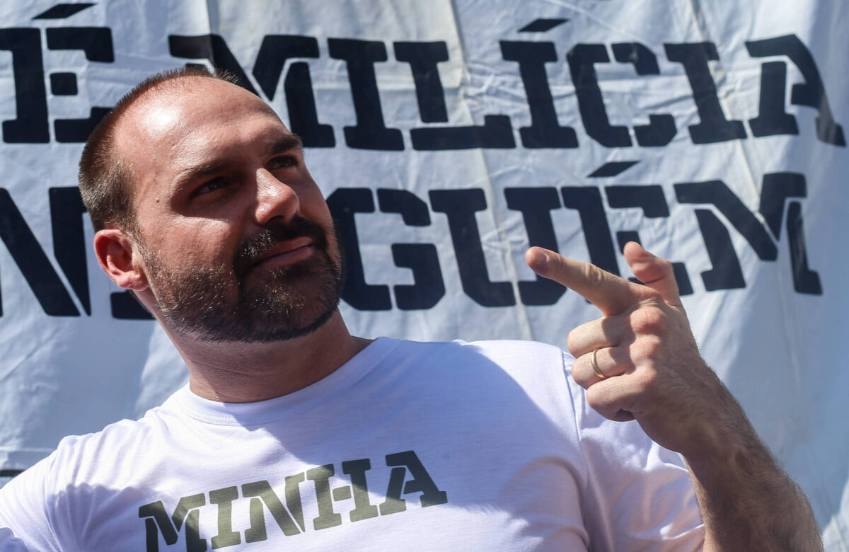 Um homem com cabelo curto e barba está posando em um evento público. Ele usa uma camiseta branca com a palavra