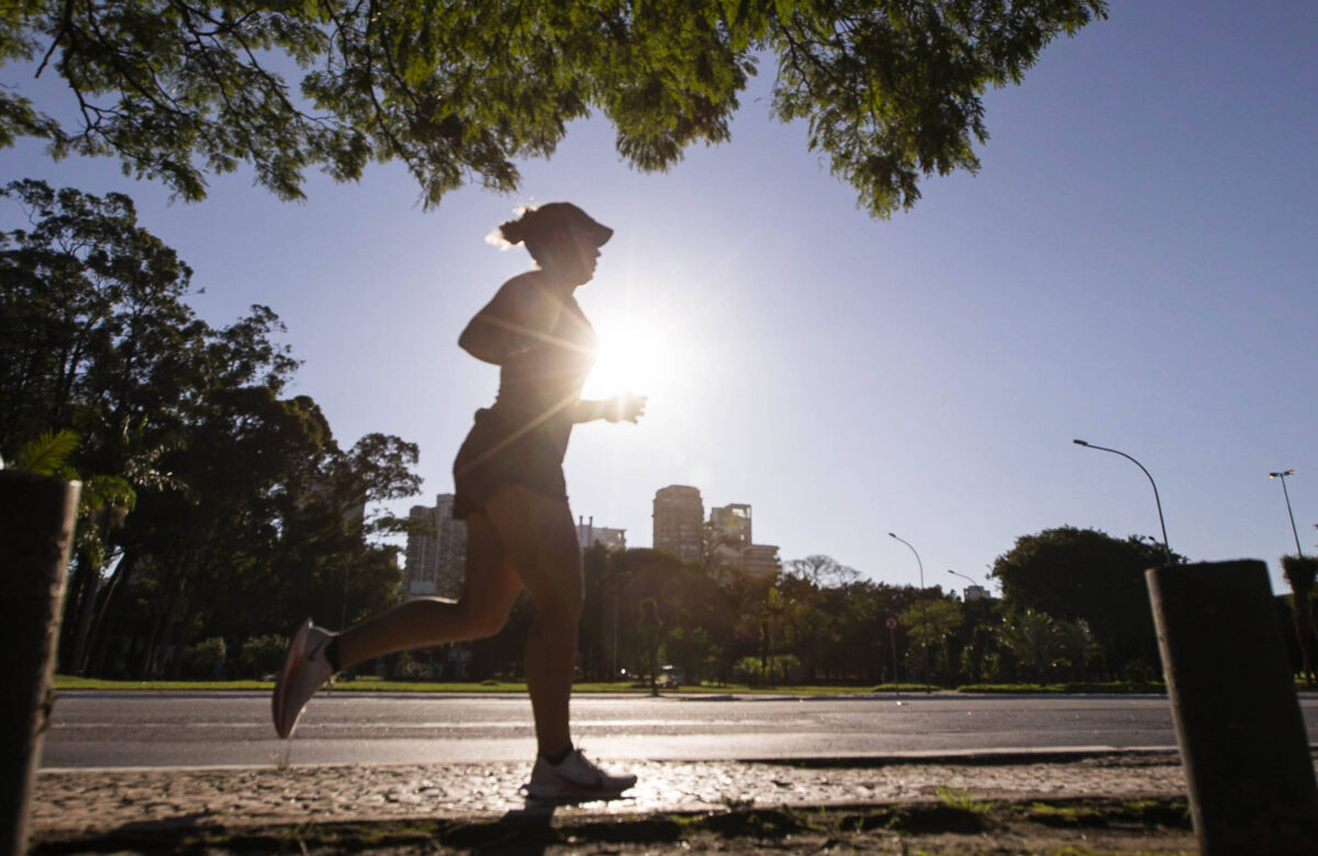 A imagem mostra uma mulher correndo em um caminho ao ar livre, com a luz do sol brilhando atrás dela, criando um efeito de silhueta. Ao fundo, é possível ver árvores e edifícios, sugerindo um ambiente urbano. O céu está claro e azul, indicando que é de manhã.