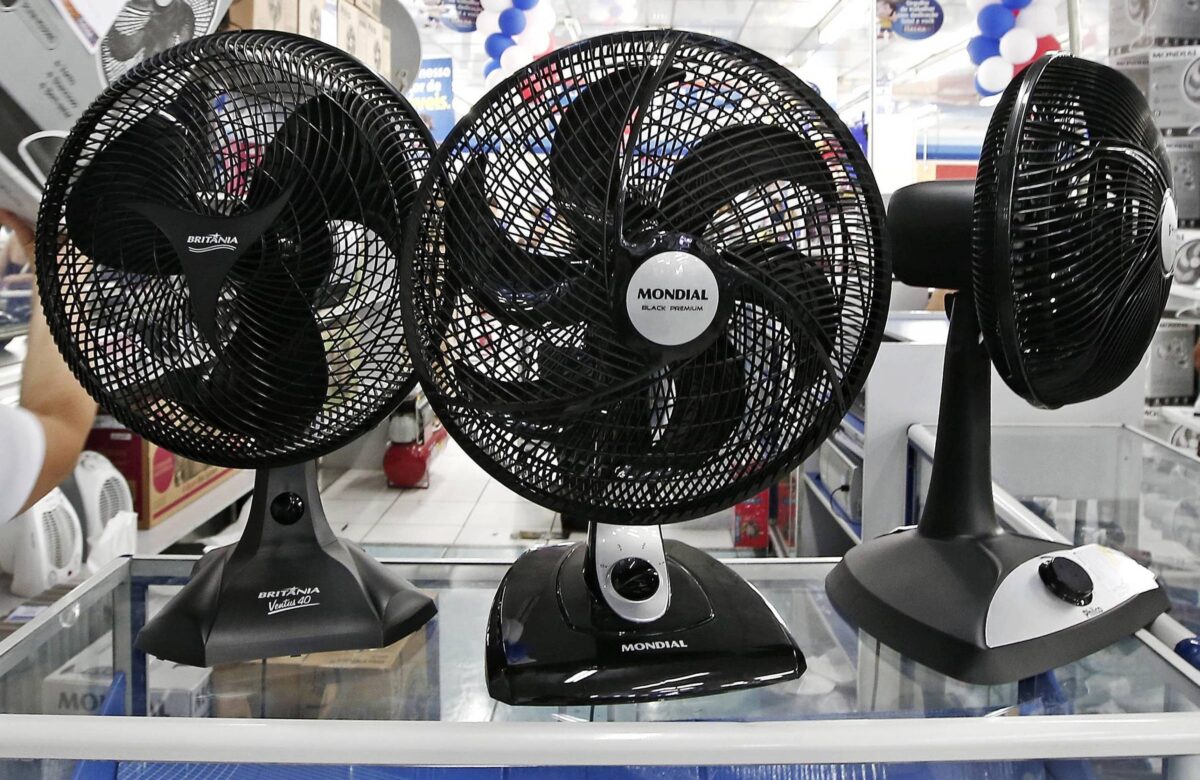 A imagem mostra uma loja com ventiladores expostos em uma prateleira. Há três ventiladores da marca Mondial em destaque, com diferentes tamanhos e modelos. Uma pessoa está segurando uma caixa, aparentemente de um ventilador, enquanto outra pessoa está ao fundo, observando os produtos. O ambiente é decorado com balões nas cores azul e branco, e há outros produtos visíveis nas prateleiras ao fundo.