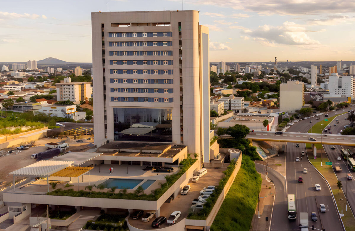 A imagem mostra um edifício moderno de vários andares, com uma fachada de vidro e um estacionamento na parte inferior. Na frente do edifício, há uma área com piscina. Ao fundo, é possível ver uma cidade com diversos prédios e uma estrada movimentada com carros. O céu está parcialmente nublado, indicando um final de tarde.