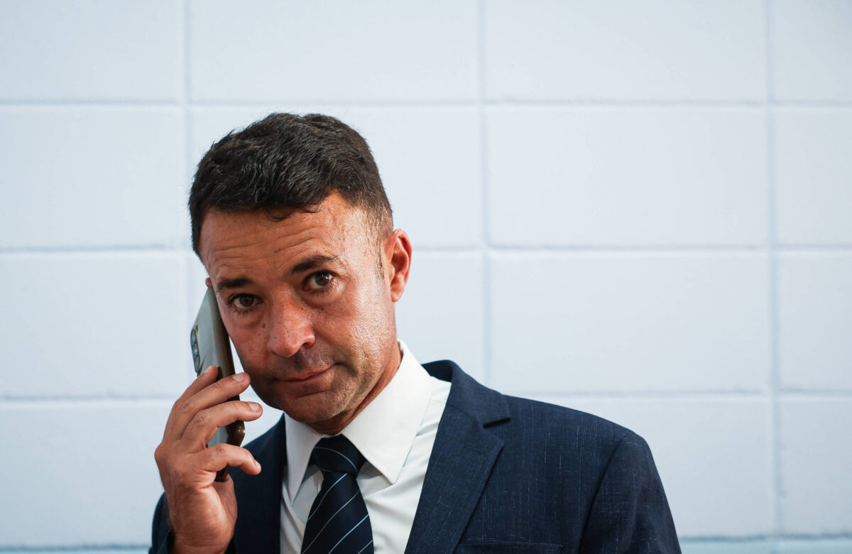 Um homem em um terno escuro está falando ao telefone. Ele tem cabelo curto e escuro, e está olhando diretamente para a câmera com uma expressão séria. O fundo é uma parede clara com azulejos brancos.