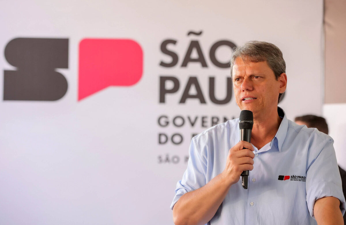 Um homem está falando ao microfone em um evento. Ele usa uma camisa azul clara e está em frente a um fundo com a logo do governo de São Paulo, que inclui as letras