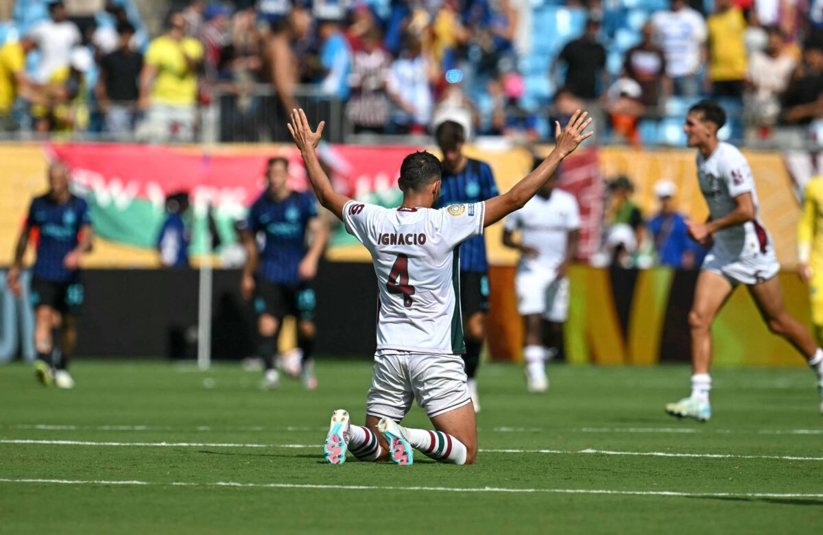 Um jogador de futebol está ajoelhado no campo, com as mãos levantadas em celebração. Ele usa um uniforme branco com detalhes em vermelho e o número 7 nas costas. Ao fundo, outros jogadores estão em movimento, e há uma multidão nas arquibancadas. O clima parece ser de comemoração após um gol.