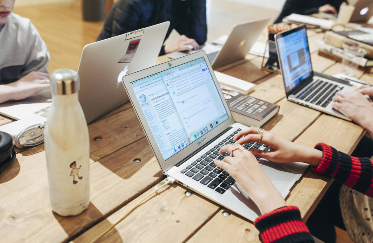 A imagem mostra uma mesa de madeira onde várias pessoas estão trabalhando em laptops. Em primeiro plano, uma mão feminina está digitando em um laptop prateado. Ao lado, há uma garrafa de água e um livro. No fundo, outros laptops estão visíveis, com algumas pessoas parcialmente visíveis. O ambiente parece ser um espaço de trabalho colaborativo.